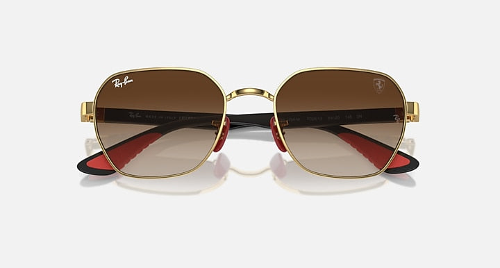 Sunglasses with gold frame and dark brown lenses, red accents near hinge, oval shape.