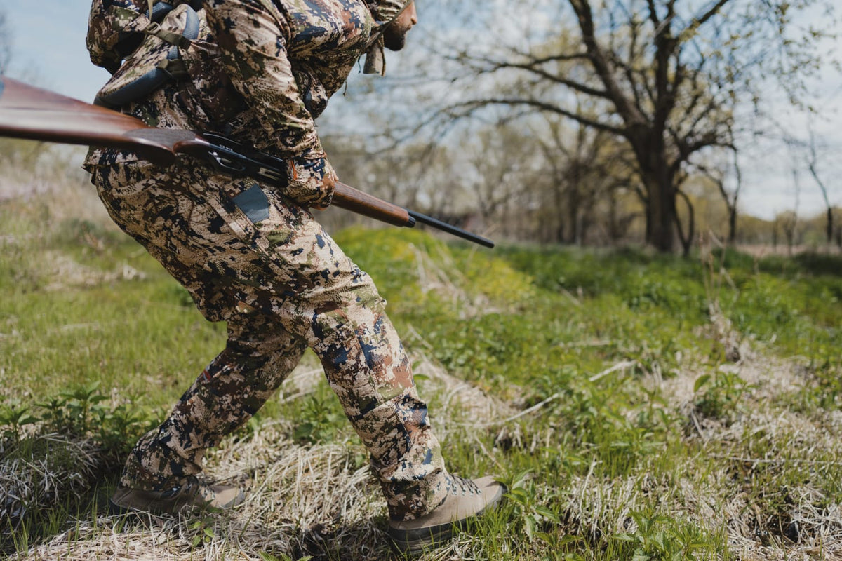 Men's camouflage hunting pants in green-brown pixel print, standing side view.