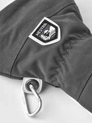 Dark gray ski glove with Hestra shield logo patch and metal ring attachment.
