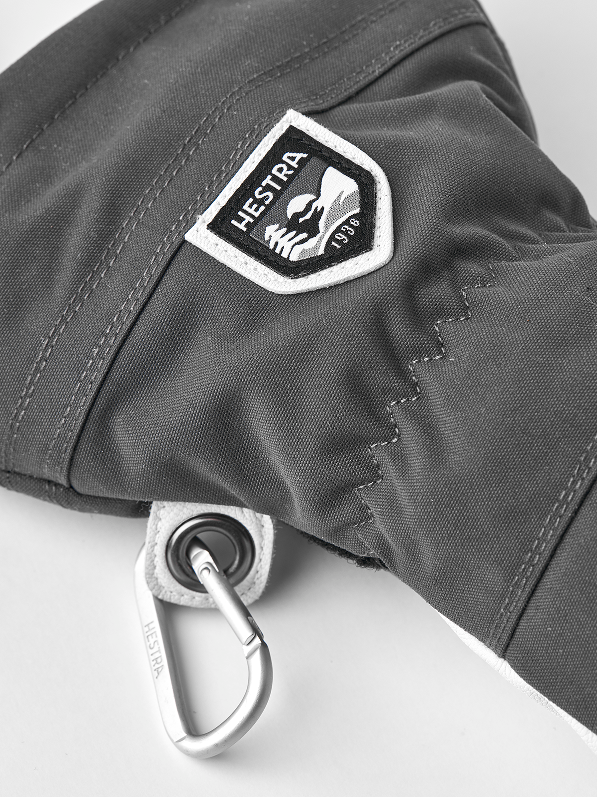 Dark gray ski glove with Hestra shield logo patch and metal ring attachment.